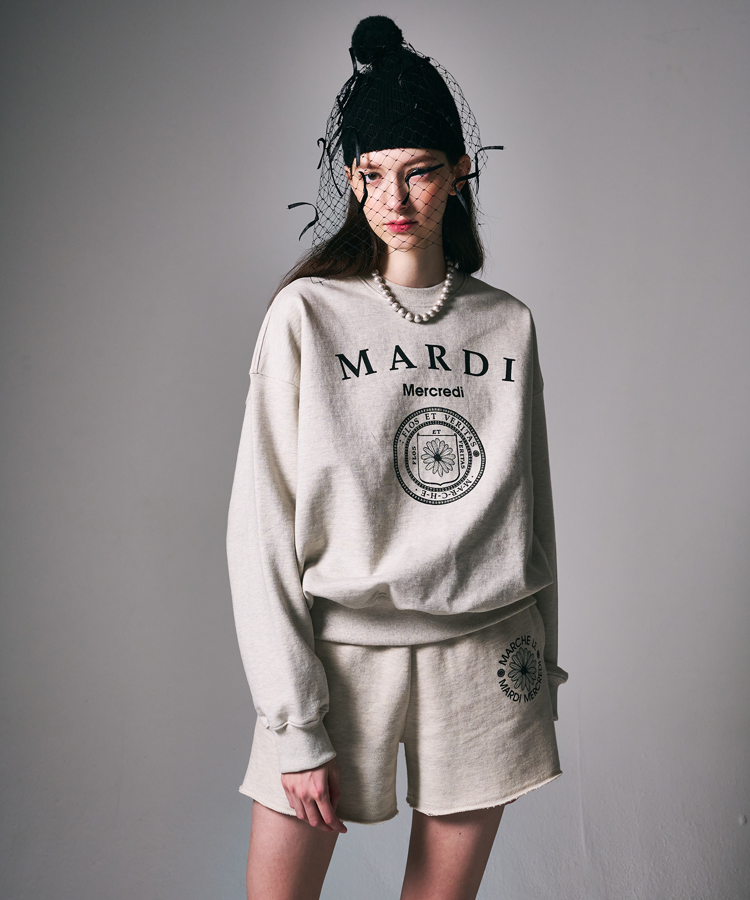 해변 야간 OX 퀴즈 장면에서 데님 팬츠와 입은 오트밀 멜란지 스웻셔츠. 가슴에 세리프 MARDI 레터링과 Mercredi 소문자, 그 아래 방패 안 데이지가 든 원형 씰 엠블럼이 블랙으로 프린트, 립 소매·밑단