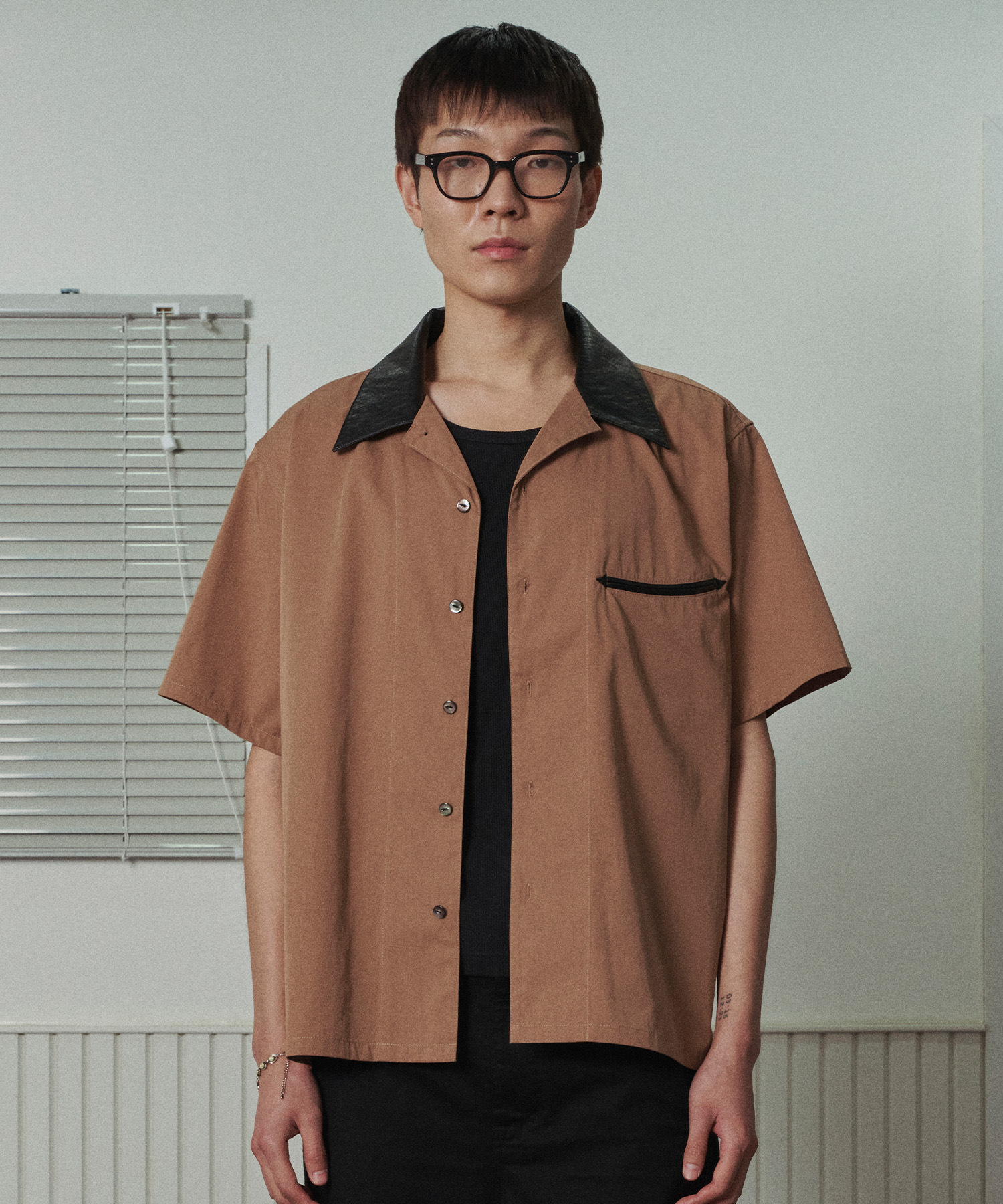 패널석에서 화이트 이너 티 위에 걸친 브라운 반팔 볼링 셔츠. 광택 있는 블랙 새틴 오픈 카라, 왼쪽 가슴에 블랙 파이핑 웰트 포켓, 앞단 다크 버튼 여밈의 박시한 핏