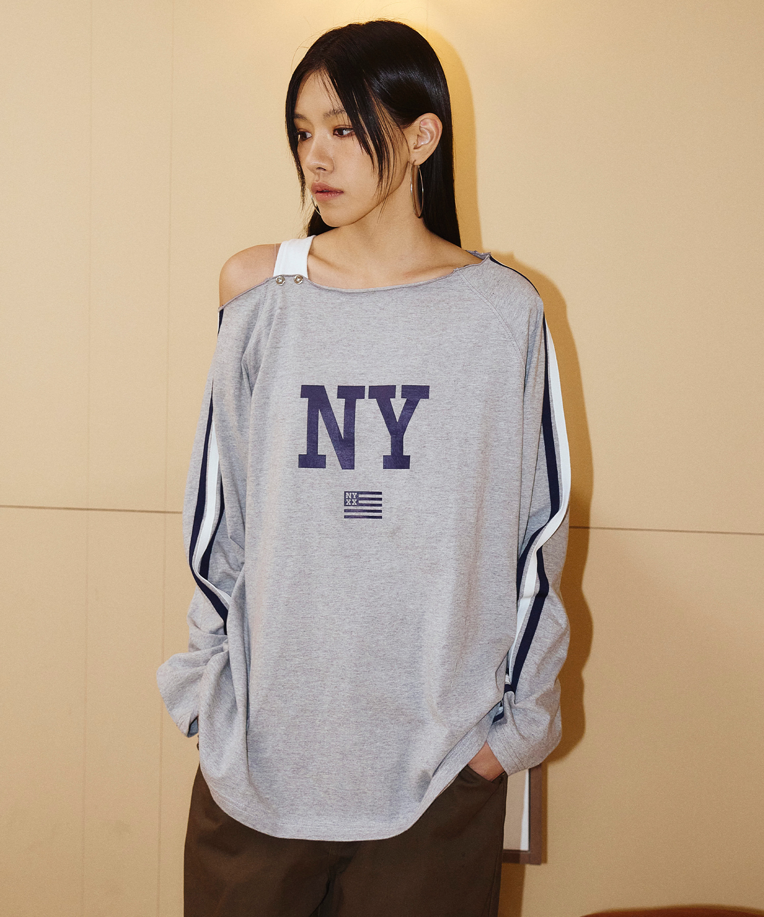 숙소 소파 장면에서 입은 그레이 멜란지 오프숄더 롱슬리브 티셔츠. 가슴 중앙 네이비 NY 레터링 아래 작은 성조기 로고, 양 소매 바깥선을 따라 네이비·화이트 이중 스트라이프 테이프, 한쪽 어깨에 스냅 단추 스트랩, 루즈핏