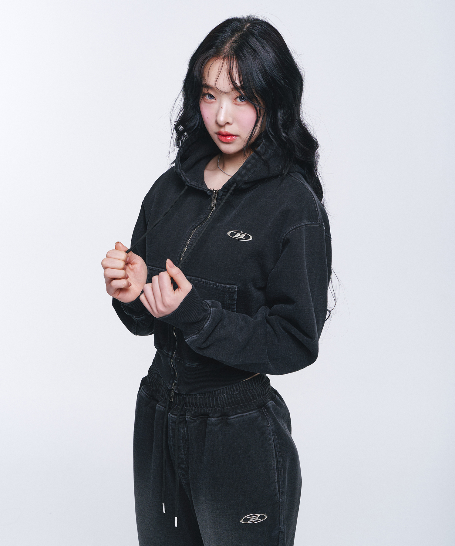 워싱된 블랙 데님 조직감의 크롭 후드 집업. 앞 양방향 지퍼, 지퍼로 갈라진 캥거루 포켓, 왼쪽 가슴에 작은 타원형 로고 자수, 소매·밑단은 립 처리에 흰 스티치가 도드라진다
