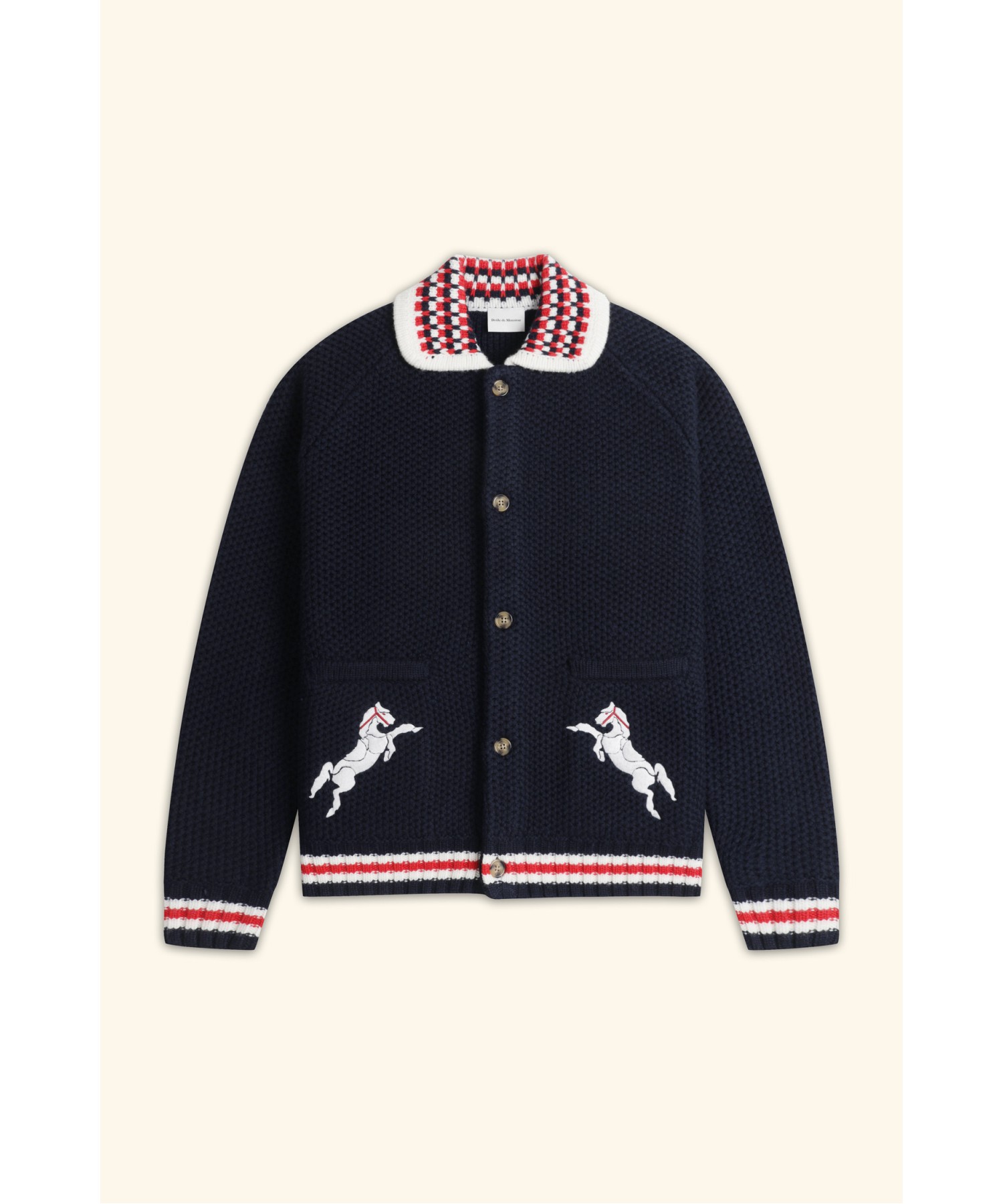 스튜디오 패널석에서 그레이 티셔츠 위에 걸친 네이비 허니콤 조직 니트 가디건. 레드·화이트·네이비 체커 니트 칼라, 소매·밑단 스트라이프 립, 양쪽 웰트 포켓 아래 화이트 말 자수, 브라운 혼 단추 5개