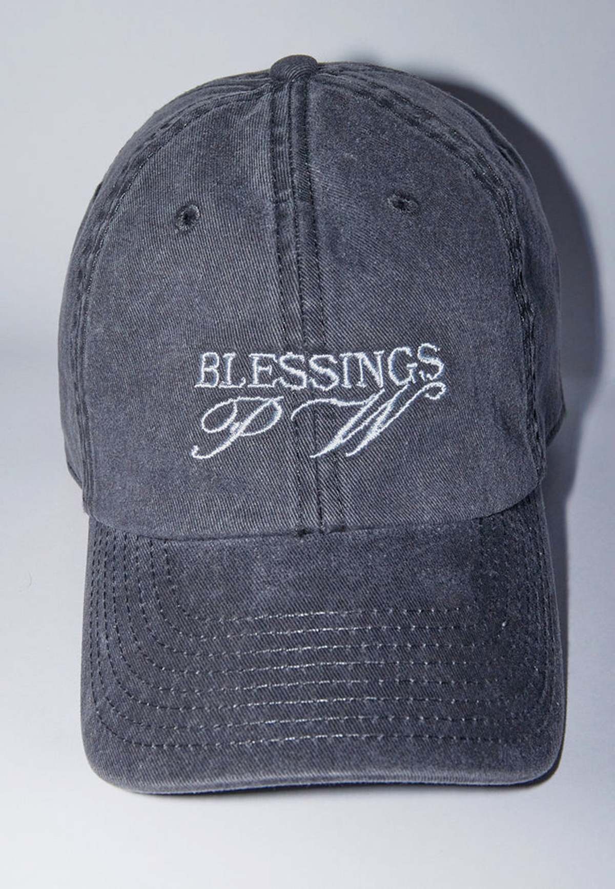 여행 중 눌러 쓴 워싱 차콜 그레이 6패널 볼캡. 정면에 흰색 'BLESSINGS' 레터링과 그 아래 필기체 이니셜 자수, 챙에 여러 줄 스티치