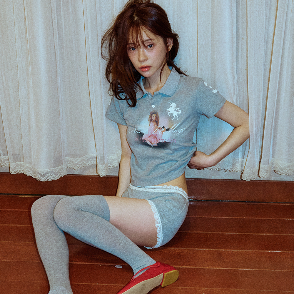 스튜디오 토크 장면에서 입은 그레이 피케 카라 티셔츠. 버튼 플래킷 아래 가슴 중앙에 유니콘과 분홍 드레스를 입은 여성을 담은 실사 그래픽과 그 아래 필기체 레터링이 프린트돼 있고, 왼쪽 소매에도 작은 화이트 프린트가 있다