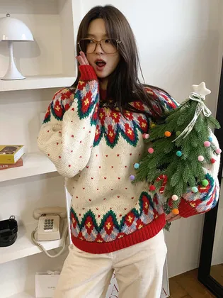 크리스마스 선물 교환 세트 장면에서 입은 아이보리 라운드넥 니트. 요크와 밑단에 레드·그린 노르딕 패턴, 레드 립 밑단
