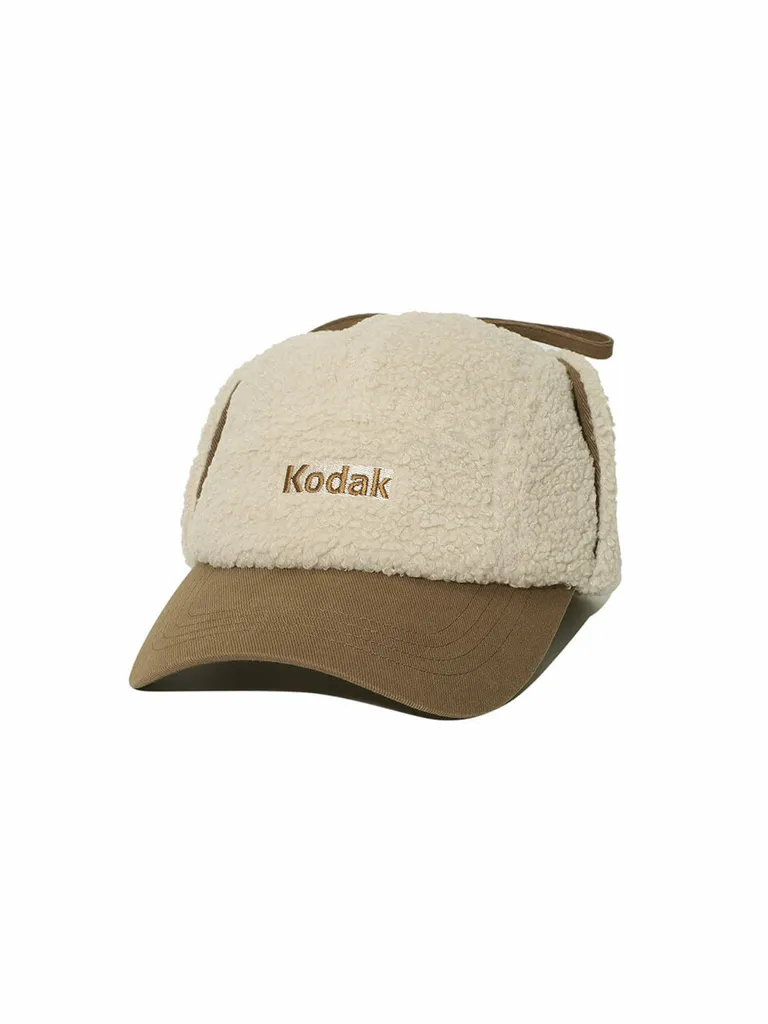 야외 장면에서 쓴 베이지 플리스 귀달이 캡. 올리브 브라운 챙과 끈, 정면 'Kodak' 자수